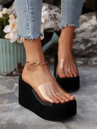 Black platform sandals with clear soles worn with light blue jeans on a stone surface.